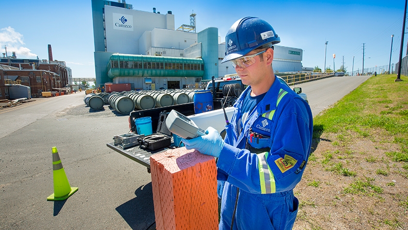 Employee wearing Cameco hard hat and coveralls looking at a handheld device in front of a Cameco building